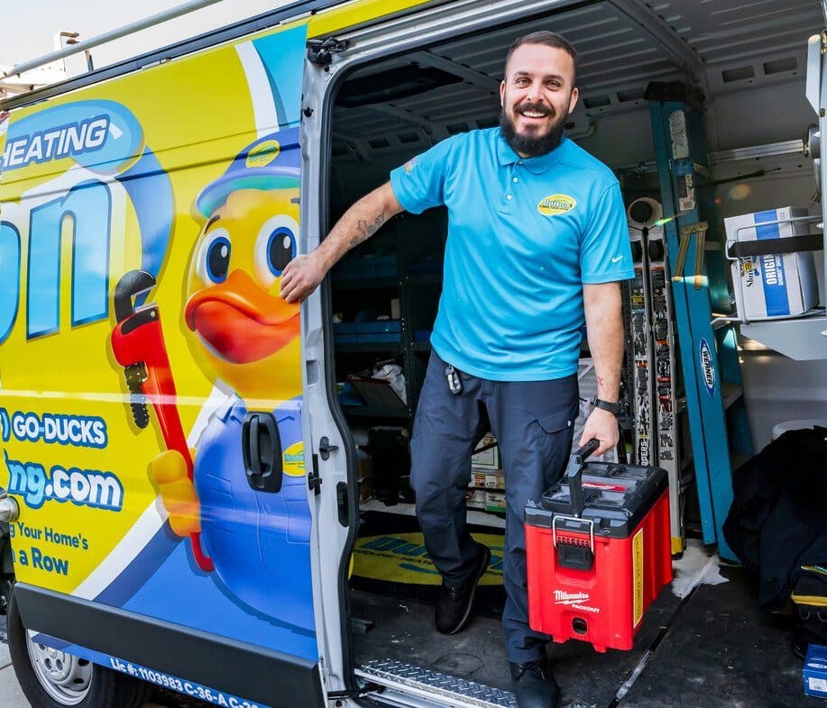 Smiling HVAC technician stepping from service van carrying a red Milwaukee toolbox beside duck-branded van graphics