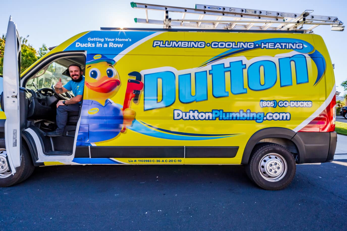 Plumber giving thumbs up from driver's seat of bright yellow Dutton Plumbing van with large duck mascot and ladder.