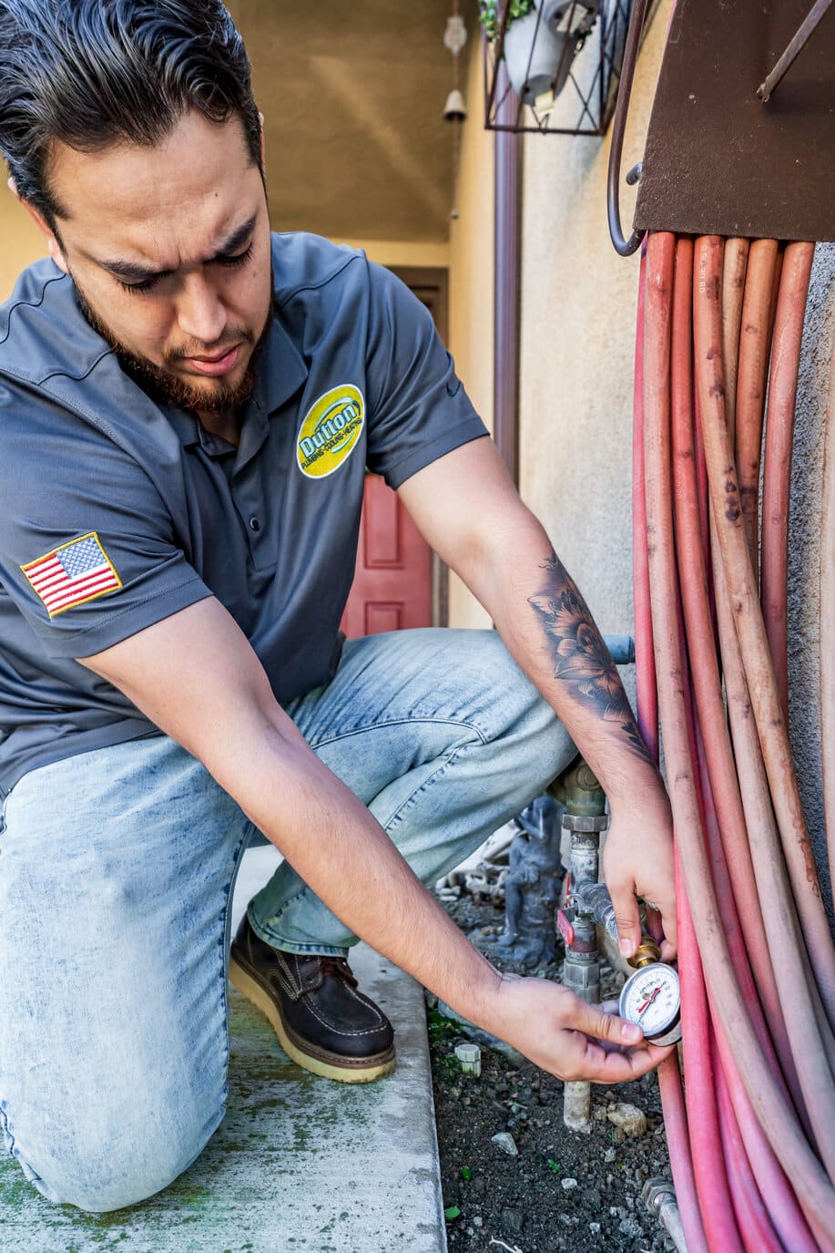 Technician kneeling outdoors checking a water pressure gauge on pipes beside coiled red hoses