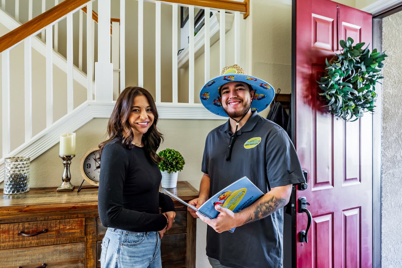 A woman and a home service technician in a colorful hat smiling and holding a brochure by a red front door.