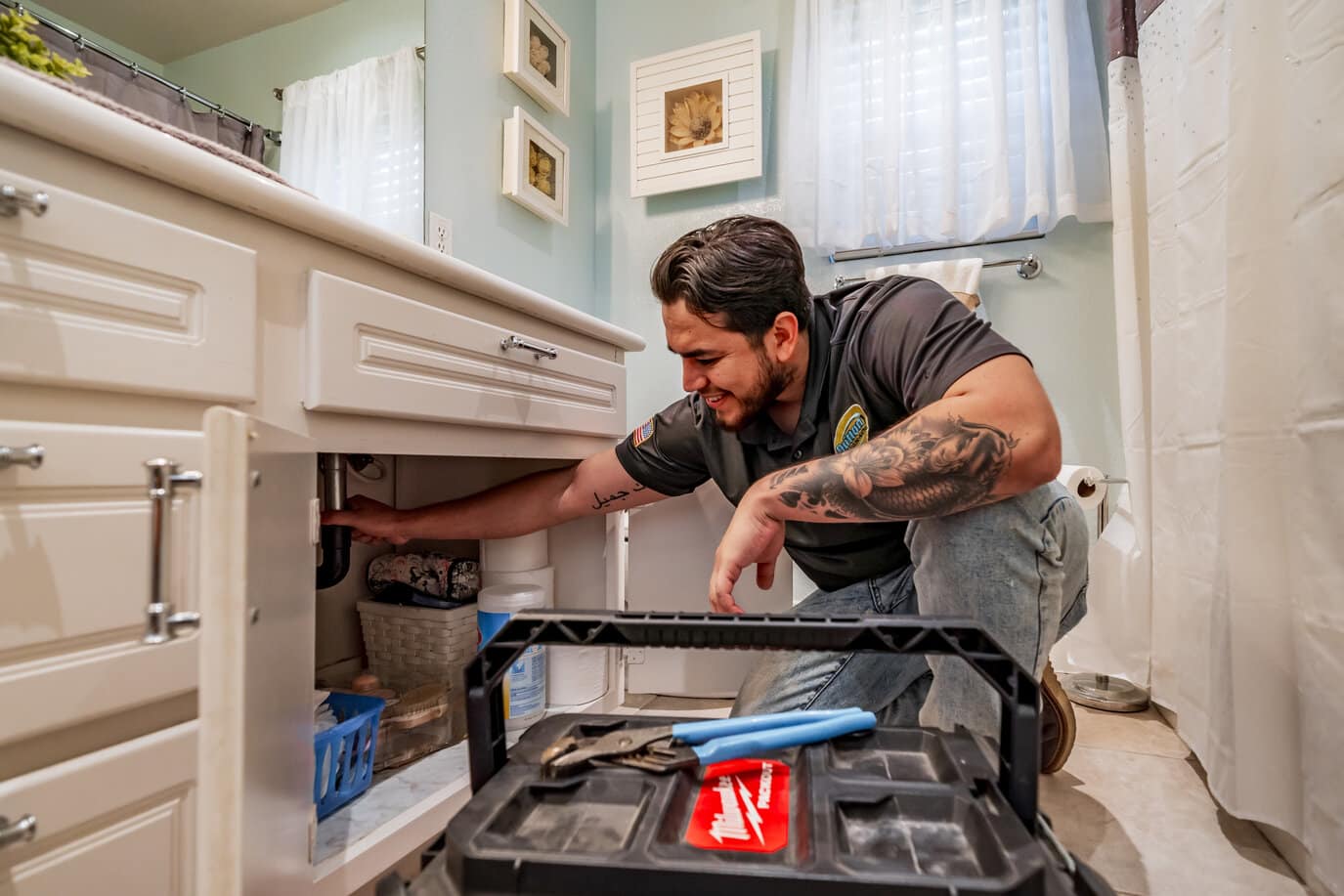 Plumber kneeling in bathroom repairing sink drain under vanity with toolbox and tools in foreground.