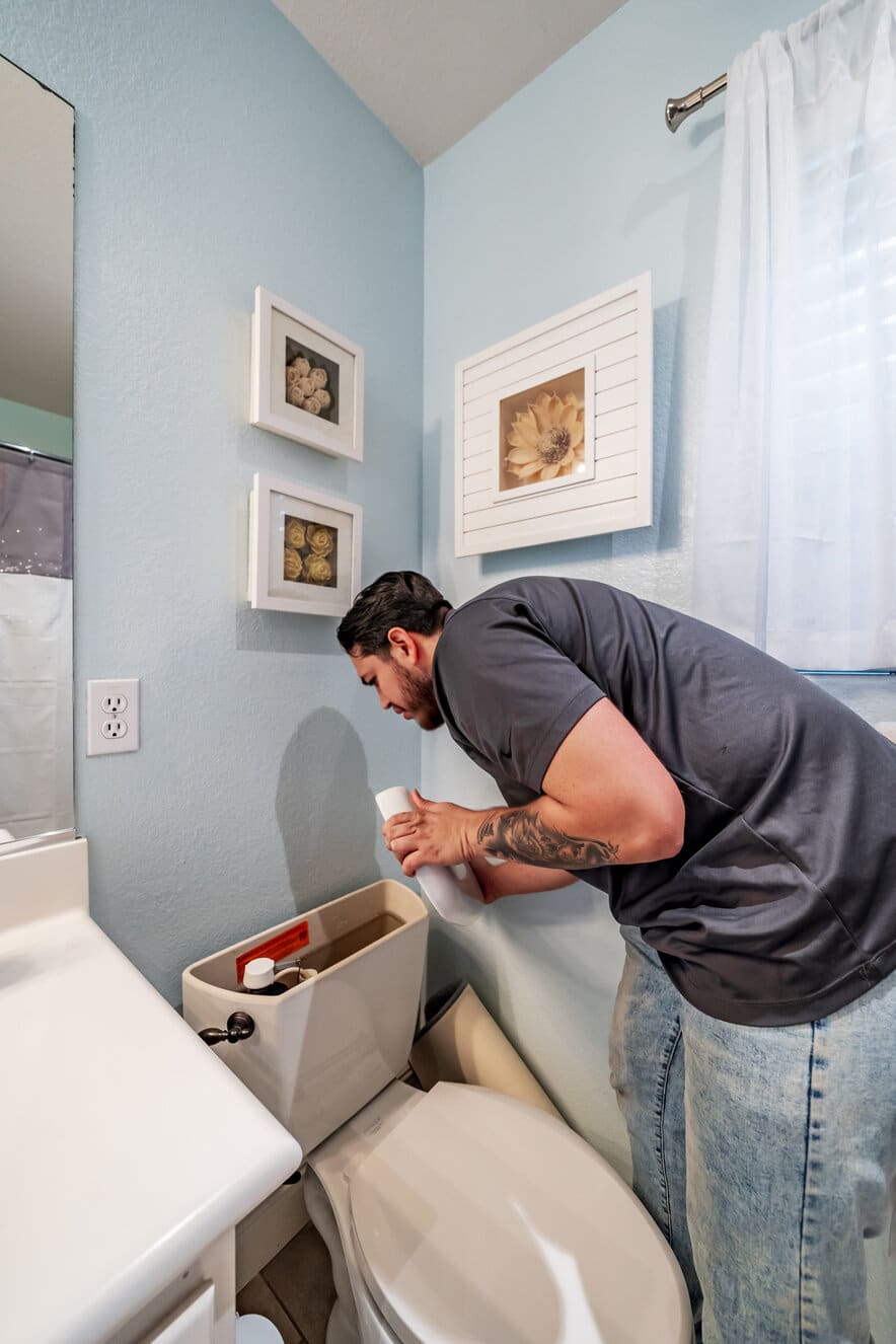 A man fixing a toilet tank in a bathroom, pouring cleaner while inspecting the flush mechanism.