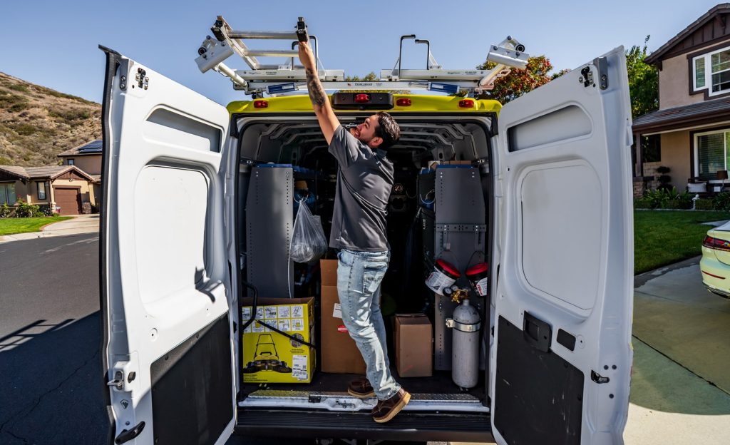 A technician reaching for a ladder on a service van with organized tools and equipment in a residential driveway.