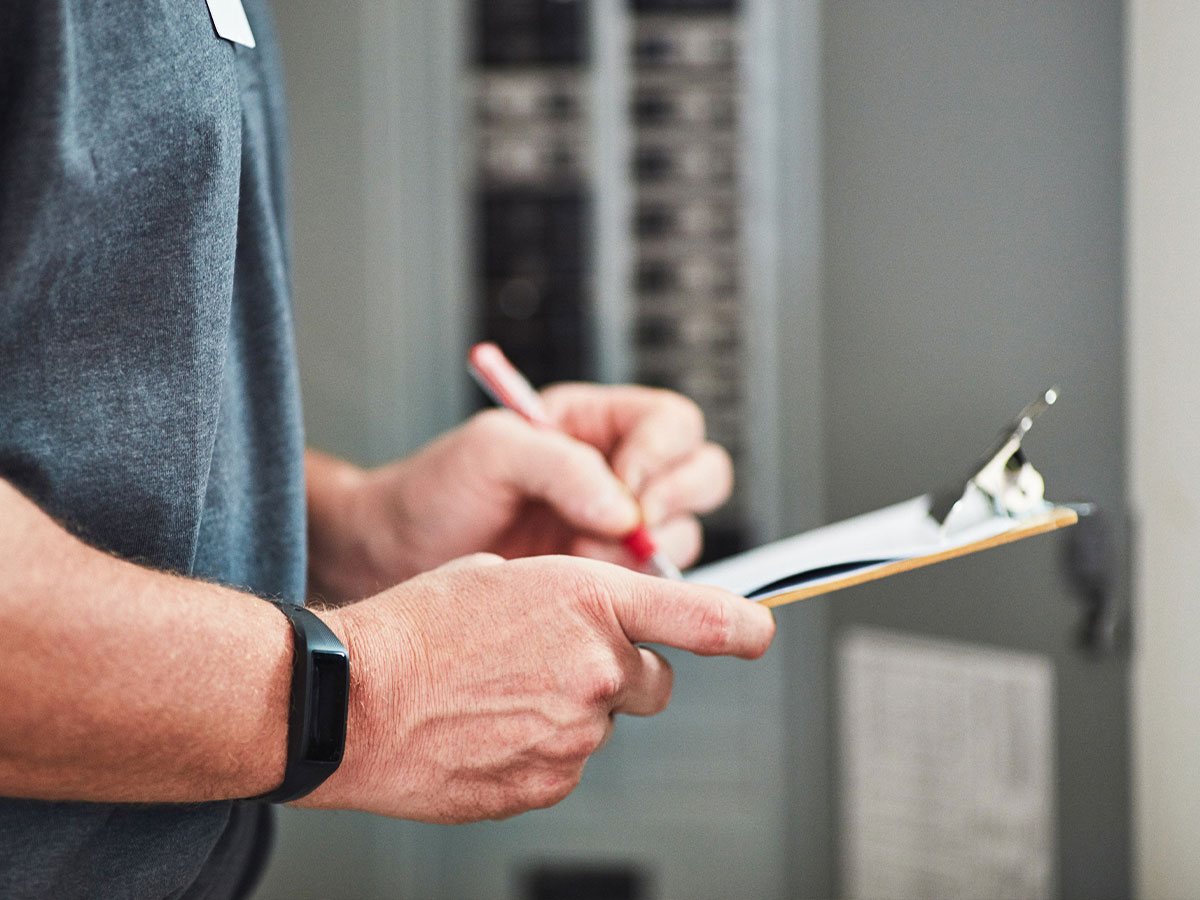 A technician's hands writing on a clipboard near an electrical panel, wearing a fitness tracker