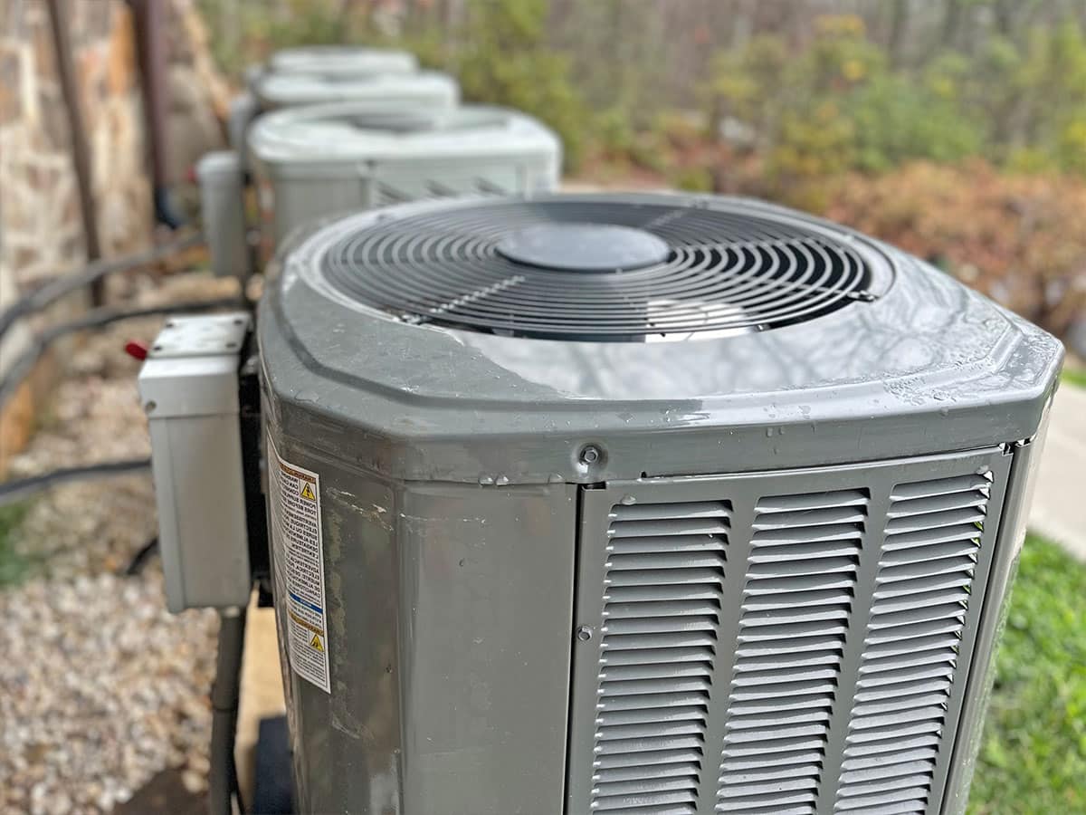 Gray outdoor air conditioning condenser unit with top fan, wet surfaces and additional units in the background
