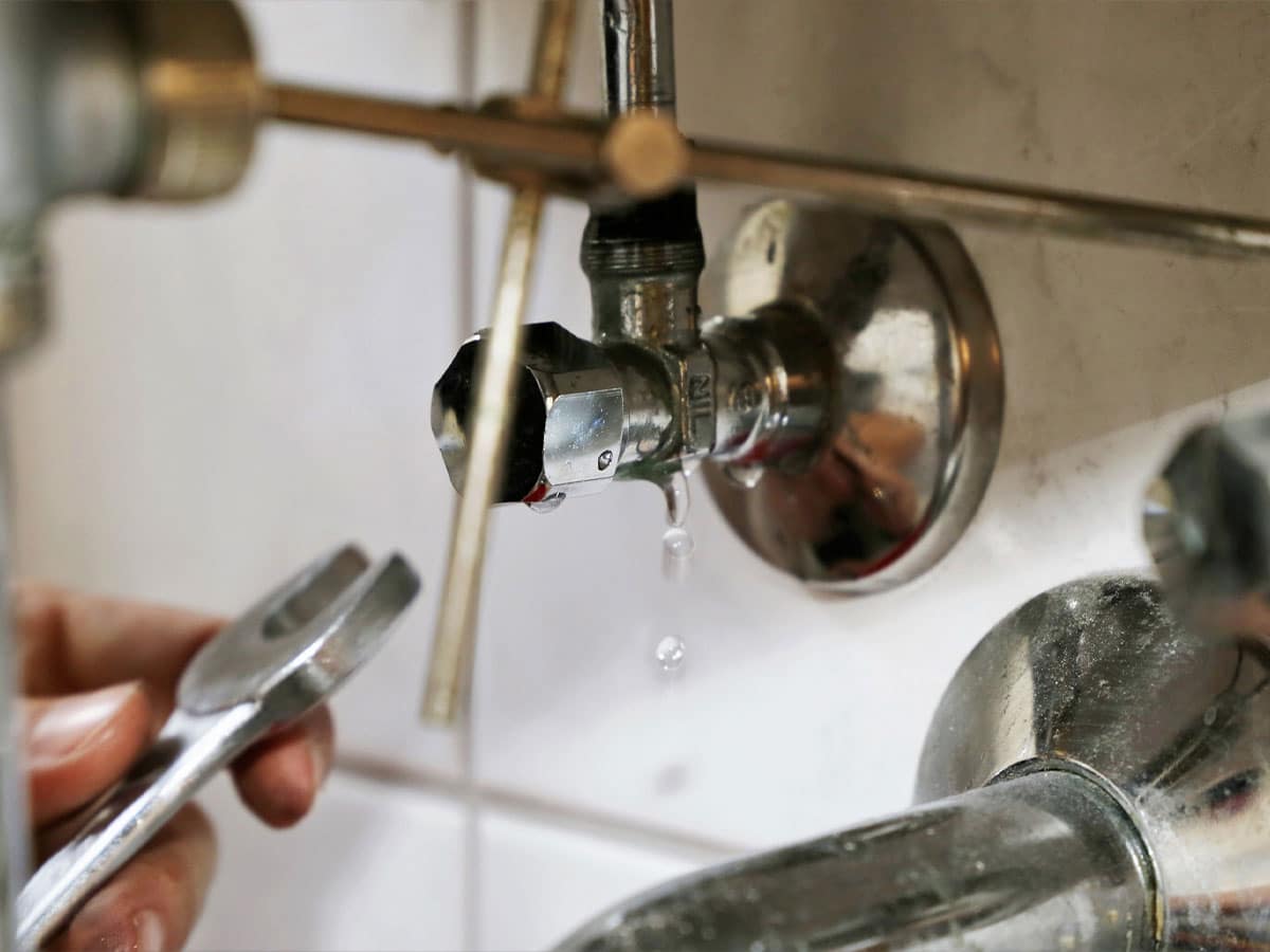 Person using wrench to tighten leaking chrome shut-off valve, water droplets falling near bathtub faucet