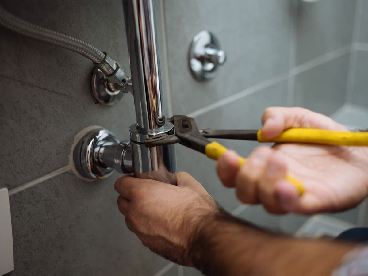 A plumber's hands using yellow-handled pliers to tighten a chrome bathroom drain pipe connection.