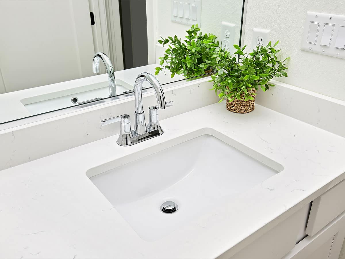 Modern white bathroom sink with chrome faucet, undermount rectangular basin, marble-look countertop and potted green plant.