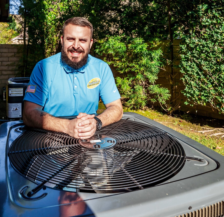 Smiling HVAC technician in blue polo leaning on an outdoor air conditioner unit in a backyard with greenery.