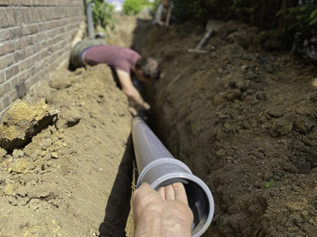 Hand guiding gray PVC drain pipe into a trench while a worker adjusts the pipe beside a brick house