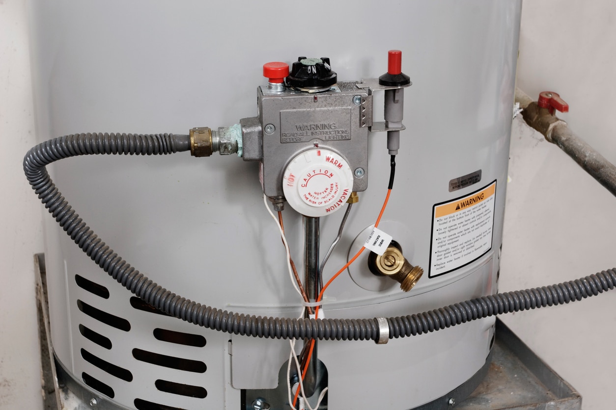 Close-up of a gray gas water heater control assembly with temperature knob, relief valve and flexible conduits