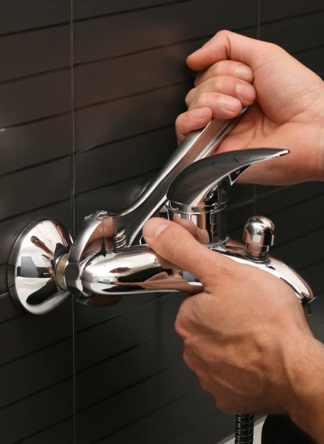Person tightening chrome shower faucet on tiled wall with adjustable wrench