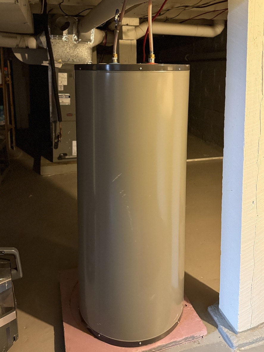 Tank-style water heater in basement on pink foam pad, copper supply lines and ductwork visible overhead