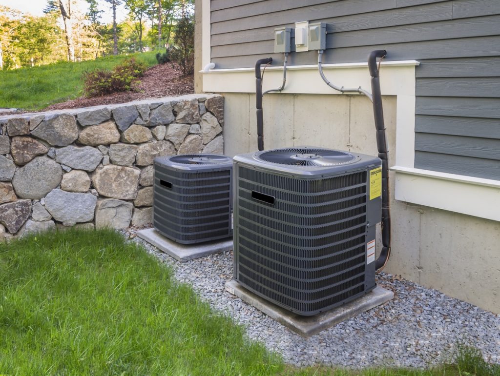 Two outdoor central air conditioning units on concrete pads beside a house with stone retaining wall and green lawn.